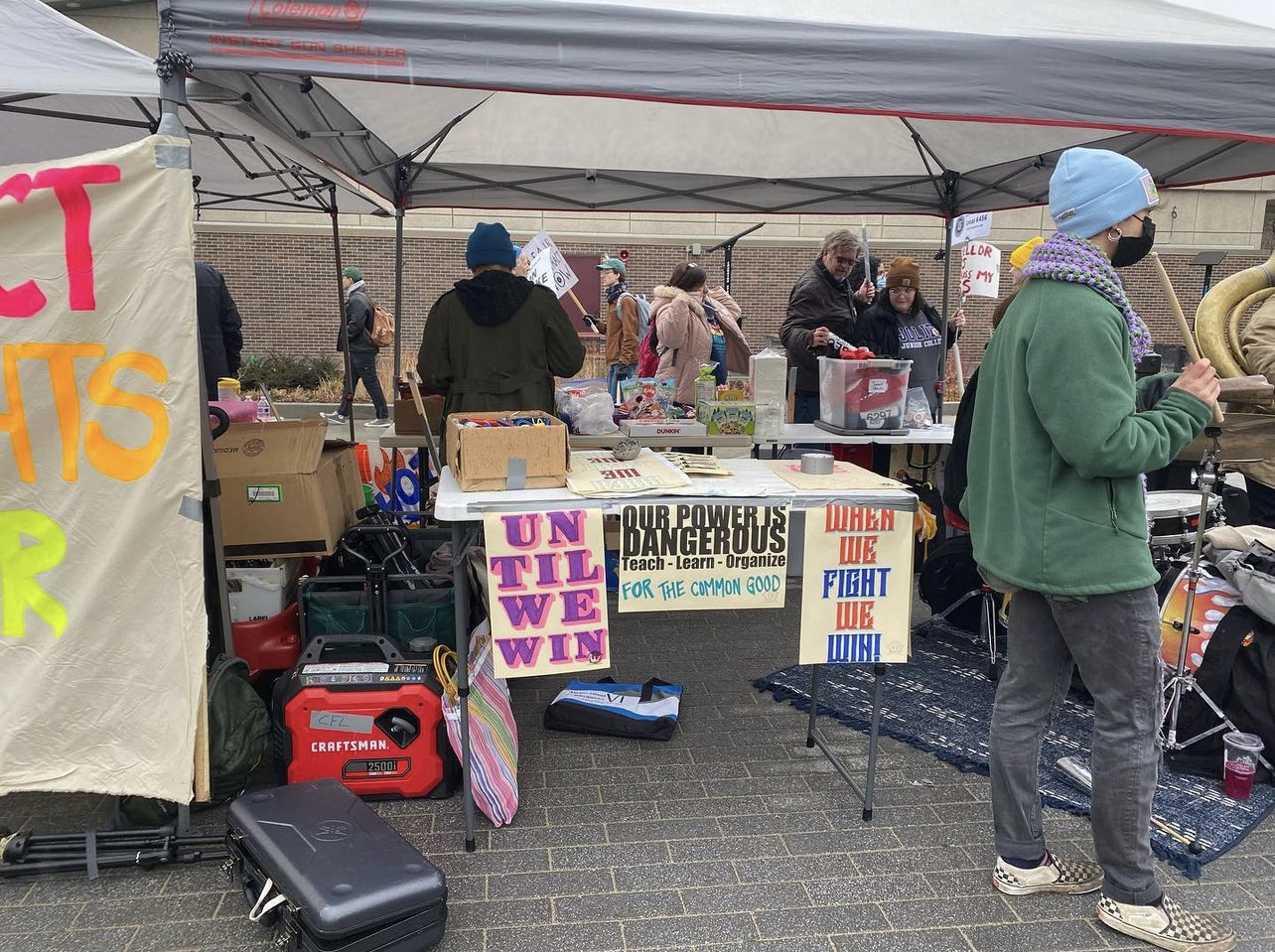 Image of a table with snacks available for anyone striking. People are standing around talking with others. There are three posters hanging form the table that read Until We Win, Our Power Is Dangerous, and When We Fight We Win.