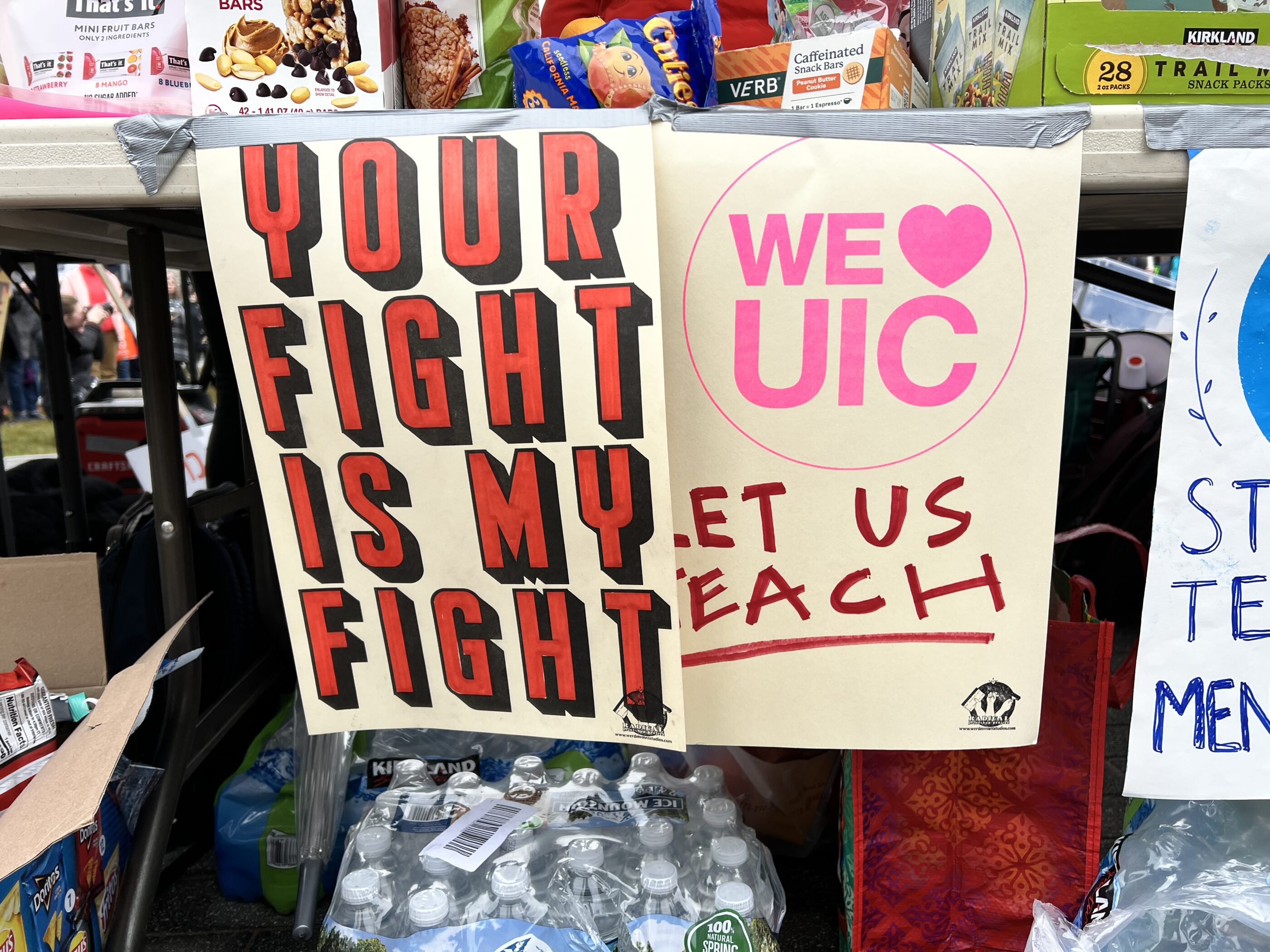Image of Posters hanging from a table. The poster on the right reads Your Fight Is My Fight and the poster on the left reads We Heart UIC Let Us Teach, with teach underlined.
